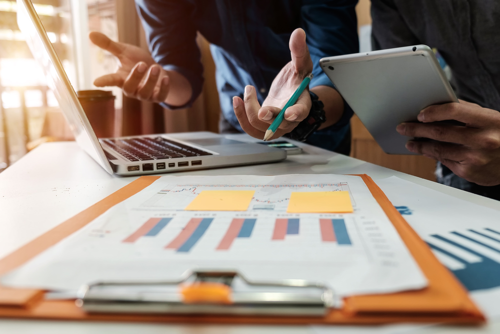  Two people are looking at a laptop and a tablet while having a discussion. A bar graph with yellow sticky notes is on the table. The image represents tips for managing finances wisely.